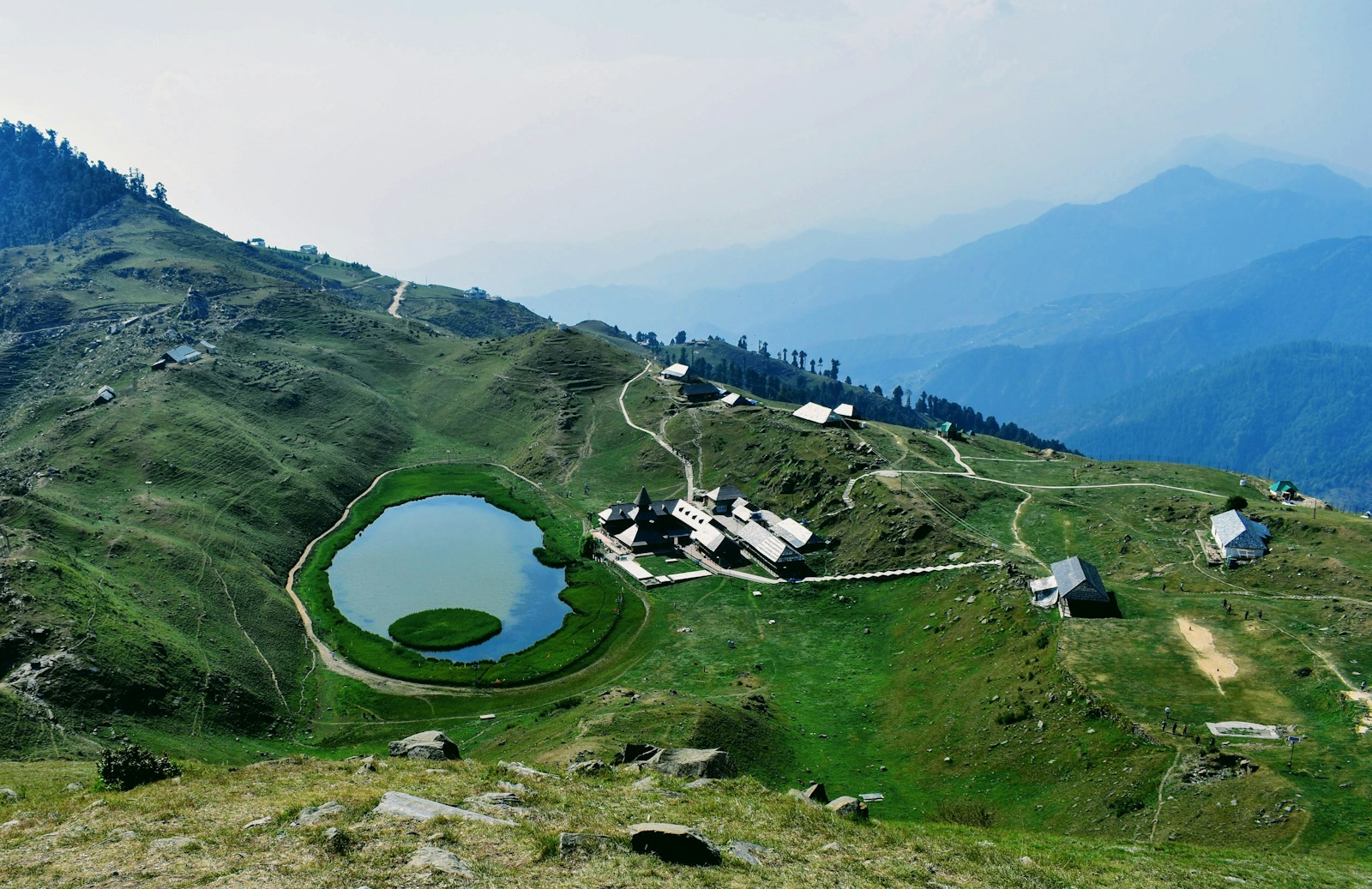 Prashar Lake and buildings amid green mountain ridges in Mandi, Himachal Pradesh.